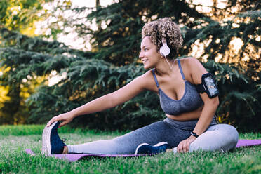 Happy young ethnic female with curly hair in sportswear listening to music with headphones and smartphone and doing seated forward bend exercise while stretching body during fitness training in summer park - ADSF13852