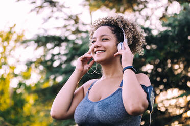 Niedriger Winkel der optimistischen jungen ethnischen Frau mit Afro-Frisur, die Kopfhörer einstellt, während sie Musik auf dem Smartphone während des Trainings im grünen Park hört - ADSF13853