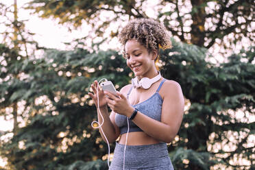 Positive young curly haired female in sportswear with headphones on neck chatting on mobile phone while resting after training in green park - ADSF13855