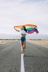 Content female with LGBT flag running along empty asphalt road and looking away - ADSF13861