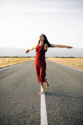 Carefree female traveler in summer dress walking along empty roadway with outstretched arms and looking away while enjoying freedom - ADSF13862