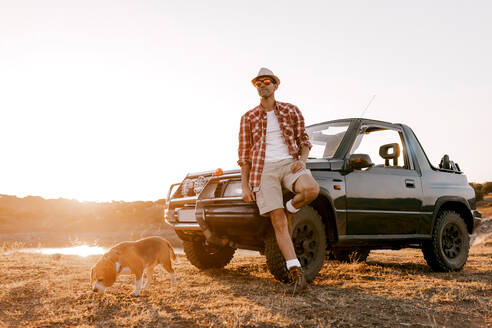 Männlicher Reisender mit Hut, der im Auto auf verblasstem Gras steht, während er mit seinem Hund unterwegs ist und die malerische Hochebene mit Bergen und Fluss im Sommer betrachtet - ADSF13890