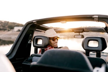 Back view of content male tourist in hat and sunglasses driving automobile near river and mountains on sunny day during vacation looking at camera - ADSF13891