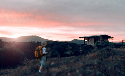 Rückenansicht eines nicht erkennbaren männlichen Reisenden mit Rucksack, der auf einem Feld in einem Bergdorf steht und den malerischen Sonnenuntergang genießt - ADSF13901