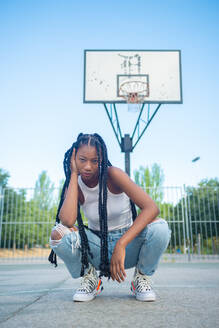 Junge Afroamerikanerin mit Zöpfen in trendigen zerrissenen Jeans und weißem Tanktop mit Turnschuhen, die auf einem Spielplatz vor einem Basketballkorb sitzt und in die Kamera schaut - ADSF13930