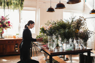 Side view of concentrated female designer arranging decorative blooming bouquets while working on order for event in creative floristry studio - ADSF13932