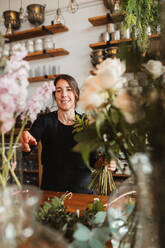 Positive junge weibliche Floristikdesignerin mit großem Strauß frischer blühender Blumen und grünem Laub an der Theke in einem gemütlichen Laden stehend - ADSF13933