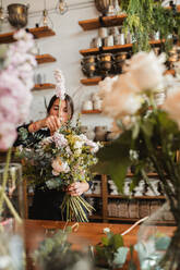 Concentrated young female floristry designer with big bouquet of fresh blooming flowers and green foliage standing at counter in cozy store - ADSF13934