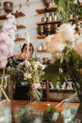 Konzentrierte junge weibliche Floristikdesignerin mit großem Strauß frischer blühender Blumen und grünem Laub an der Theke in einem gemütlichen Laden stehend - ADSF13934