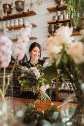Positive young female floristry designer with big bouquet of fresh blooming flowers and green foliage standing at counter in cozy store - ADSF13935