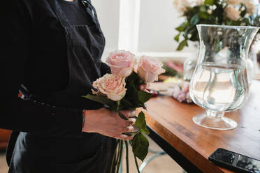 Seitenansicht einer nicht erkennbaren Floristin, die schöne zartrosa Rosen hält, während sie an einem Tresen mit einer mit Wasser gefüllten Glasvase steht, während sie in einem Floristiksalon arbeitet - ADSF13936