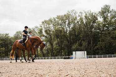 Ebenerdige Darstellung von weiblichen Jockeys in Uniform, die in Sätteln sitzen und Pferde auf einer sandigen Koppel während des Trainings reiten - ADSF13947