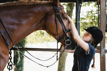 Side view of busy female equestrian in helmet standing in stable and preparing horse for training while putting bridle - ADSF13961