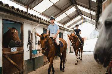 Ernste weibliche Jockeys in Helmen und Stiefeln reiten auf Kastanienpferden entlang der Scheune auf einer Ranch und schauen weg - ADSF13969