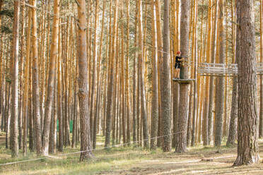 Kind in Sicherheitsausrüstung, das auf einer hölzernen Plattform auf einem Baum steht und sich im Abenteuerpark im Wald amüsiert - ADSF13990