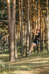 Cheerful boy in protective helmet and safety harness riding zip line and having fun in adventure park in summer - ADSF13991