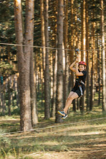 Fröhlicher Junge mit Schutzhelm und Sicherheitsgurt, der mit einer Seilrutsche reitet und im Sommer im Abenteuerpark Spaß hat - ADSF13991