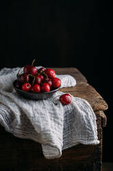 Bow full of fresh cherries on wooden table and white muslin fabric against dark background - ADSF13996
