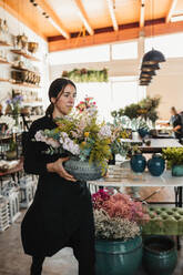 Professionelle Floristin in schwarzer Uniform, die einen Keramiktopf mit einem Strauß blühender Blumen und Grünpflanzen trägt, während sie in einem Floristikstudio arbeitet - ADSF14016