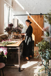 Seitenansicht einer konzentrierten Designerin, die dekorative blühende Sträuße arrangiert, während sie an einem Auftrag für eine Veranstaltung in einem kreativen Floristikstudio arbeitet - ADSF14017