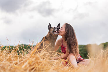 Frau im Sonnenkleid mit geschlossenen Augen streichelt sanft reinrassigen Hund mit glattem Fell, während in Feld mit verblasstem Gras in der Nähe von Wald in der Landschaft unter Himmel mit Wolken sitzen - ADSF14065