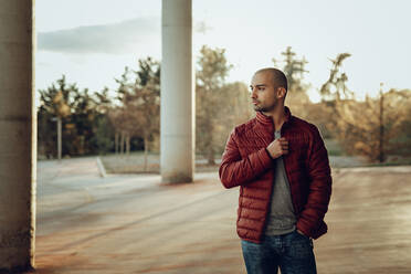 Confident young unshaven male in red warm jacket and jeans looking away while standing on paved square against blurred trees in sunny autumn day - ADSF14093