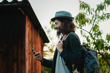 Seitenansicht eines männlichen Hipsters mit Rucksack, der in einer ländlichen Gegend steht und auf seinem Mobiltelefon surft - ADSF14095