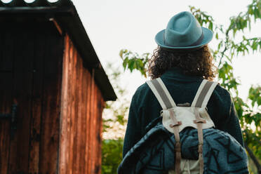 Von hinten Blick auf anonyme Mann Hipster mit langen Haaren und Rucksack stehen in der Landschaft neben einem hölzernen Hütte im Sommerurlaub - ADSF14096