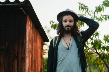 Zufriedener männlicher Hipster mit langen Haaren und Rucksack, der im Sommerurlaub auf dem Land steht und in die Kamera schaut - ADSF14097