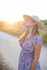 Charmante Frau in stilvollem Kleid und Strohhut stehen auf sandigen Straße in ländlicher Umgebung und genießen Sonnenuntergang am Wochenende - ADSF14130