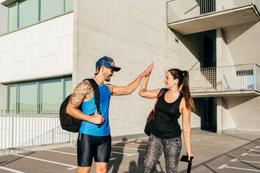 Smiling couple of athletes in sportswear standing on street and giving high five after training while looking at each other - ADSF14132