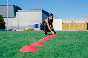 Junge sportliche Frau in Sportkleidung ordnet Trainingskegel auf grünem Rasen an, während sie sich für Beweglichkeitsübungen im Stadion vorbereitet - ADSF14139