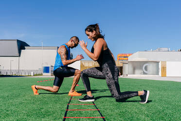 Full body side view of focused sporty man and woman doing lunges while exercising with agility ladder on green grassy lawn on sports ground - ADSF14144