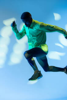 Ganzkörper-Seitenansicht eines jungen afroamerikanischen männlichen Läufers in einem bunten Trainingsanzug, der in einem Studio mit heller Neonbeleuchtung aus der Startposition sprintet - ADSF14153
