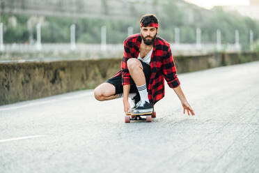 Männlicher Hipster in kariertem Hemd, der auf einem Longboard sitzt und die Straße entlang fährt - ADSF14165