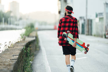 Rückenansicht eines männlichen Skaters in kariertem Hemd und mit Longboard, der an einem Sommerwochenende in der Stadt auf einer asphaltierten Straße spazieren geht - ADSF14166