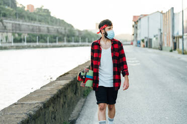 Young male skater in surgical mask and with longboard walking along promenade during coronavirus pandemic and looking away - ADSF14167