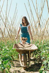 Happy girl and mother walking near wheelbarrow in garden - ADSF14173
