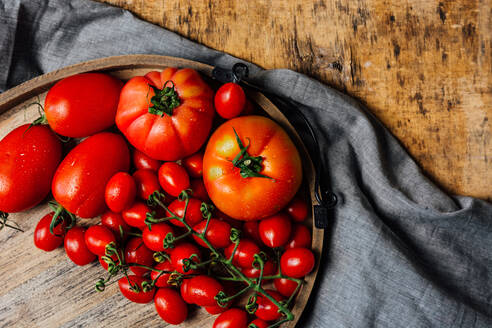Draufsicht auf verschiedene Sorten frischer reifer roter Tomaten auf einem Holztablett auf einem rustikalen Holztisch mit Tuch - ADSF14184