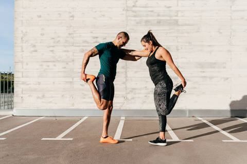 Side view of strong sportsman and sportswoman leaning on each other and stretching legs before training in city on sunny day stock photo