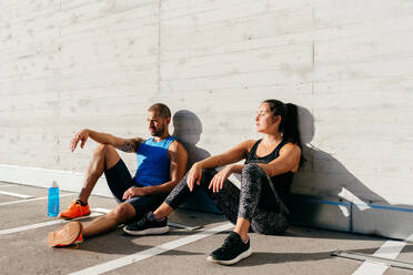 Couple of exhausted athletes in sportswear sitting on street and leaning on stone wall while resting after intense workout on sunny day in city - ADSF14189