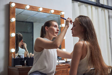 Positive junge weibliche Make-up-Künstlerin macht Kunden Gesicht Make-up mit Pinsel in der Nähe von gemütlichen großen Spiegel mit Lichtern in zeitgenössischen Schönheitssalon - ADSF14192