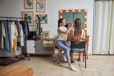 Positive junge weibliche Make-up-Künstlerin macht Kunden Gesicht Make-up mit Pinsel in der Nähe von gemütlichen großen Spiegel mit Lichtern in zeitgenössischen Schönheitssalon - ADSF14193