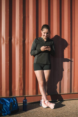 Frau lehnt sich an einen Container und benutzt ein Smartphone, lizenzfreies Stockfoto