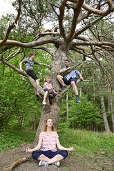 Vater und Kinder sitzen auf einem Ast, während die Frau unter einem Baum im Wald meditiert - ECPF01026
