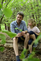 Lächelnder Vater im Gespräch mit seiner Tochter, während er auf einem Baumstamm im Wald sitzt - ECPF01037