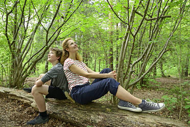 Thoughtful mother and son sitting back to back on fallen tree in forest - ECPF01039