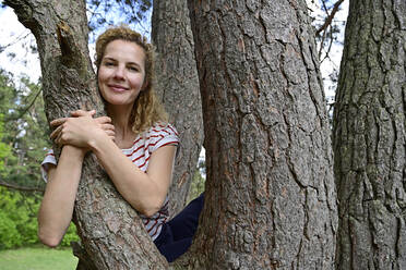 Lächelnde Frau, die sich umarmt, während sie auf einem Baum im Wald sitzt - ECPF01042
