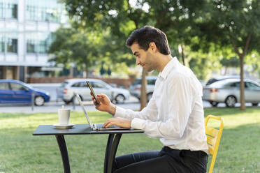 Geschäftsmann, der ein Mobiltelefon und einen Laptop benutzt, während er in einem Straßencafé sitzt - AFVF07129