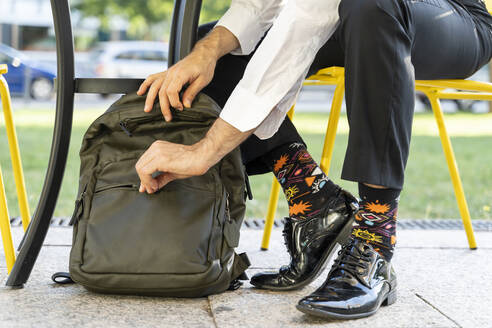 Männlicher Unternehmer mit bunten Socken, der seinen Rucksack schließt, während er auf einem Stuhl in einem Straßencafé sitzt - AFVF07141